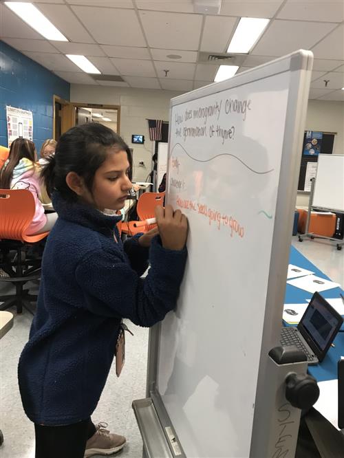 Girls writes notes on dry erase board while students seated in background work in groups. 